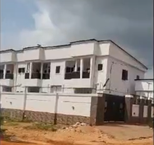 Modern white apartment building with balconies, perimeter fencing, and gated entrance in a developing residential neighborhood.
